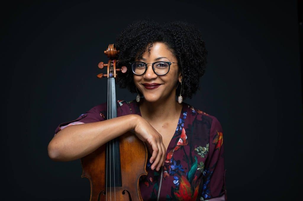 Ashe Gordon, a Black woman in a floral top wearing glasses, smiles at the camera. She holds a violin under her arm.