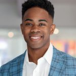 A black man wearing a light blue, plaid suit jacket over a white button-down shirt. He has a short, tapered haircut and is smiling at the camera.