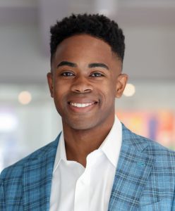 A black man wearing a light blue, plaid suit jacket over a white button-down shirt. He has a short, tapered haircut and is smiling at the camera.