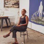 A photo of a black person, wearing a black tank top and black jeans sitting on a chair. They have their left leg crossed over their right, their hands are folded on their lap, and she is smiling. They are wearing a necklace and have their hair, locs, tied back. She is sitting in a studio with white walls. There is a blue fiber art piece behind them to the left.