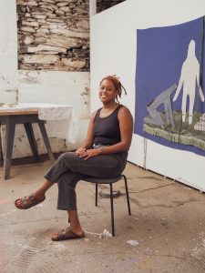 A photo of a black person, wearing a black tank top and black jeans sitting on a chair. They have their left leg crossed over their right, their hands are folded on their lap, and she is smiling. They are wearing a necklace and have their hair, locs, tied back. She is sitting in a studio with white walls. There is a blue fiber art piece behind them to the left.