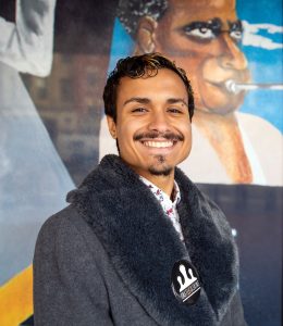 A young man smiling at the camera. They have tan skin and short brown hair that curls at the front. He is wearing a grey jacket with a dark grey fur collar. On the collar is a round pin that says "Castle of Our Skins". Their left eyebrow is pierced along with his septum and right nostril. Behind him is a mural of someone playing the trumpet.