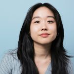 Portrait style of photo of a young Asian American woman. Her hair is black, long and parted down the middle. She is facing the camera and smiling. She is wearing small gold hoops and a baby blue cardigan. The background is also blue.