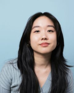 Portrait style of photo of a young Asian American woman. Her hair is black, long and parted down the middle. She is facing the camera and smiling. She is wearing small gold hoops and a baby blue cardigan. The background is also blue.