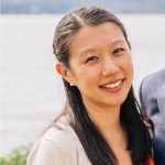 Photo of young Asian American woman smiling at camera. There is a body of water behind her. Her hair is long, black, parted in the middle and tucked behind her ear. It falls past her shoulders. She is wearing a cream colored shirt,