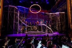 The cast of Carousel on a stage set with a roller coaster, with the orchestra in the foreground